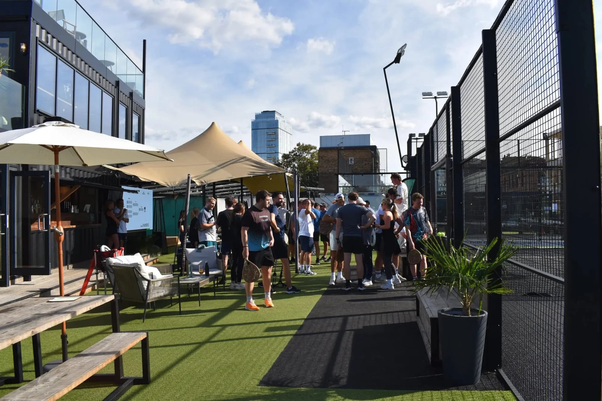 People gathered on an outdoor padel court with umbrellas and seating