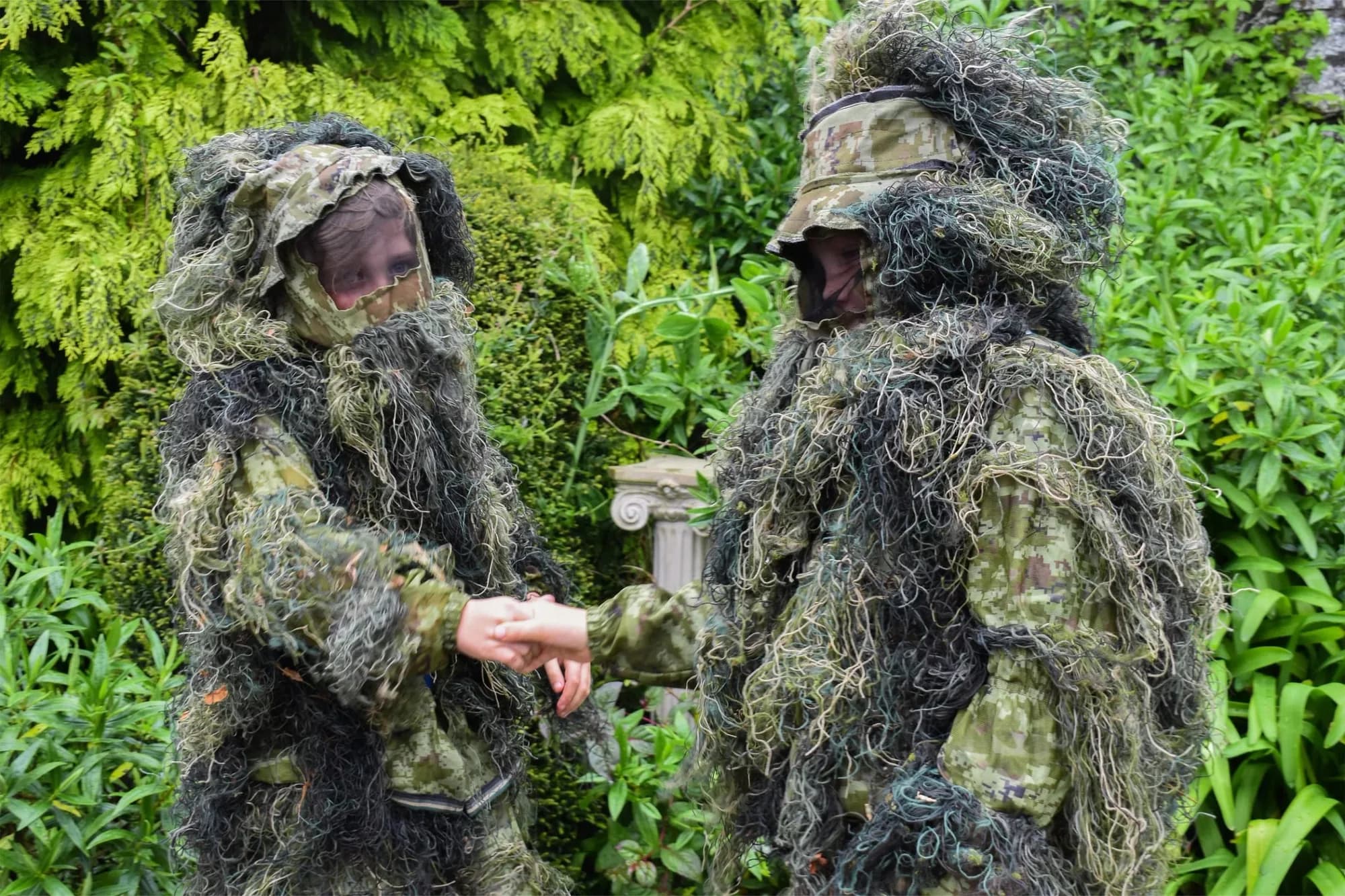 Two people in camouflage and ghillie suits shaking hands in the woods
