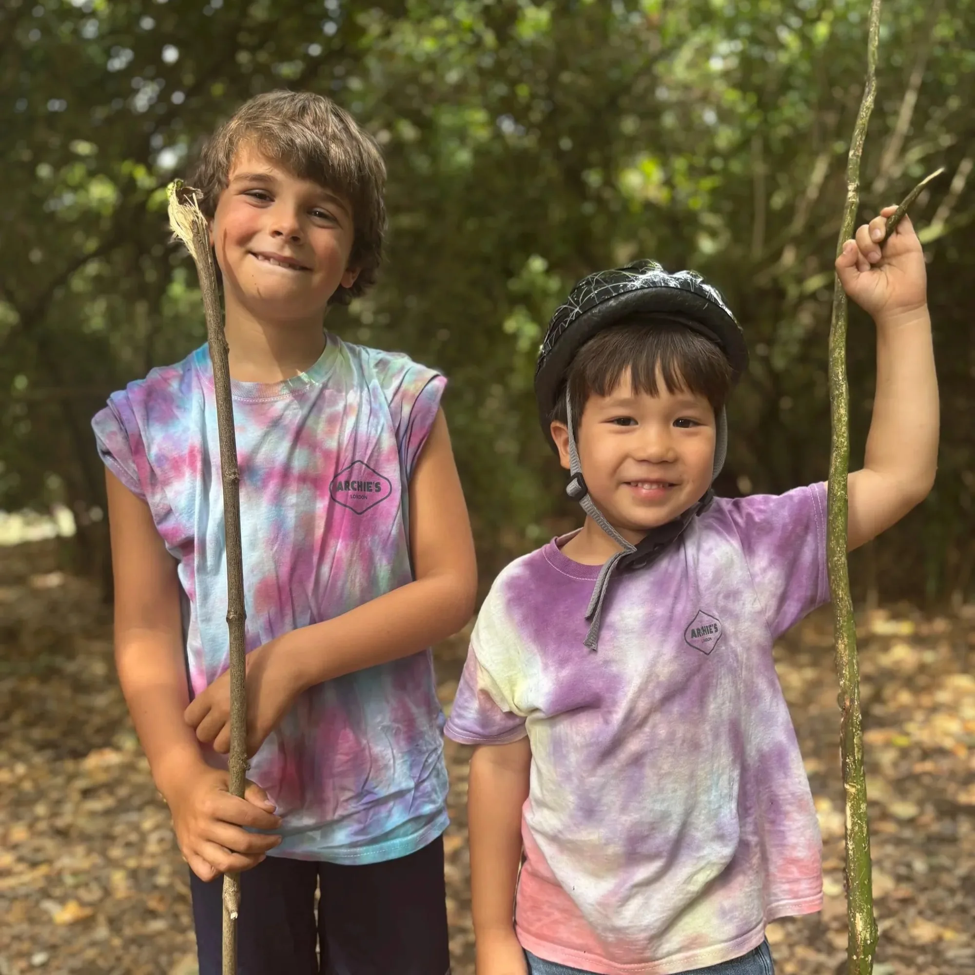 Two young boys outdoors holding sticks with a wooded background